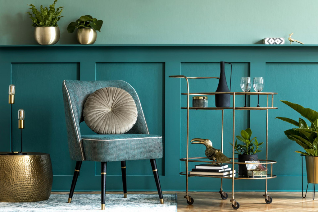 Dizajn interijera luksuzne dnevne sobe u boji petrolej-zelene sa stilskom foteljom, zlatnim kolicima za piće, biljakama i elegantnim osobnim dodacima. Zelena je i zidna obloga s policom