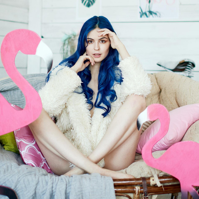girl with long legs and blue hair in a fur jacket posing in a chair with a pink flamingo in a vintage studio