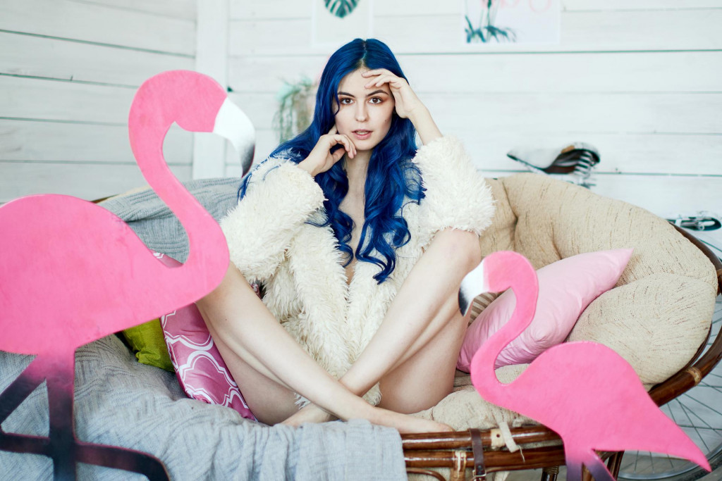 girl with long legs and blue hair in a fur jacket posing in a chair with a pink flamingo in a vintage studio