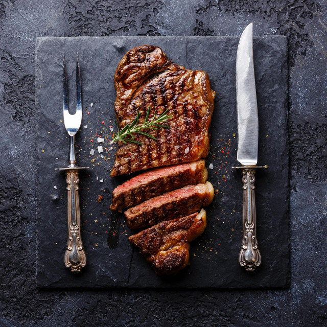 Sliced grilled meat barbecue steak Striploin with knife and fork carving set on black stone slate