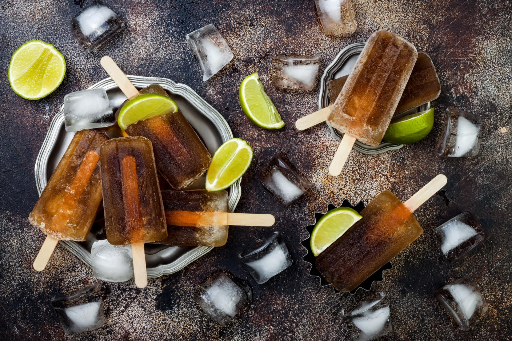 Rum and coke cocktail popsicles with lime juice. Cuba libre homemade frozen alcoholic paletas - ice pops. Overhead, flat lay, top view, copy space