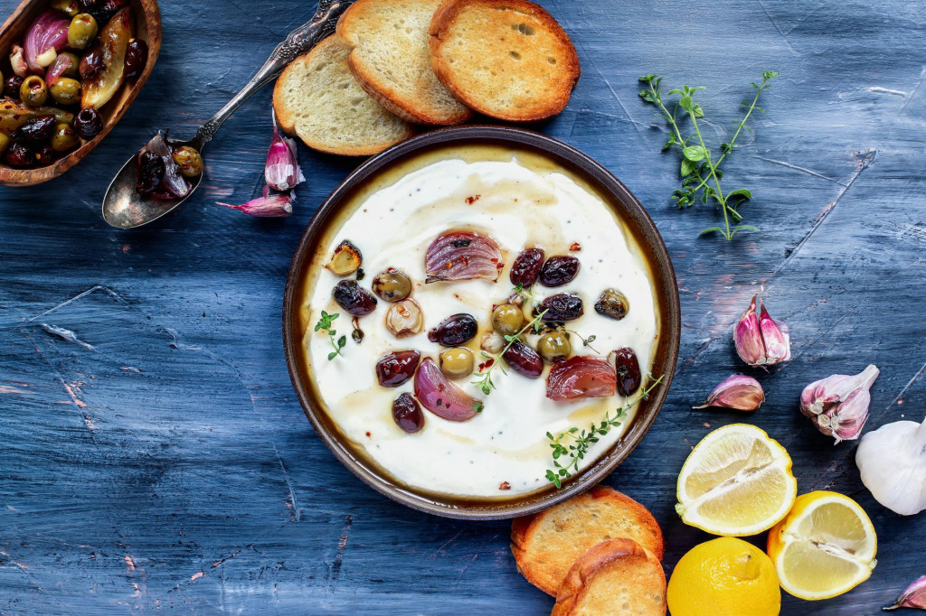 Flatlay of Greek feta cheese dip, Tirokafteri, with roasted olives, Italian garlic, red onions or shallots, drizzled with olive oil and garnished with lemon thyme. Top down view.