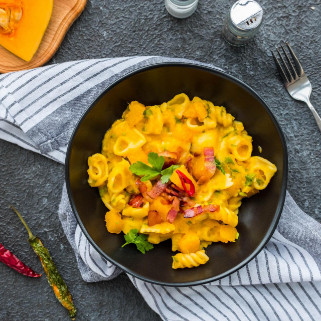 Pasta with pumpkin, fried bacon and hot peppers in a black plate on a dark concrete background. Pasta recipes. Italian food.