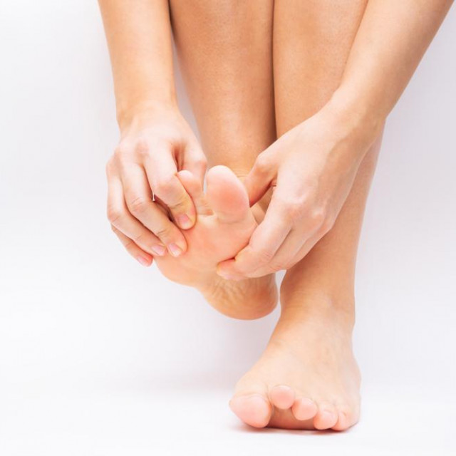 Pain in the foot and toes from uncomfortable shoes. A woman holds a sore foot with her hands on a white background. Pathology of bone structures, flat feet. Cramp, convulsion, spasm. Orthodontics