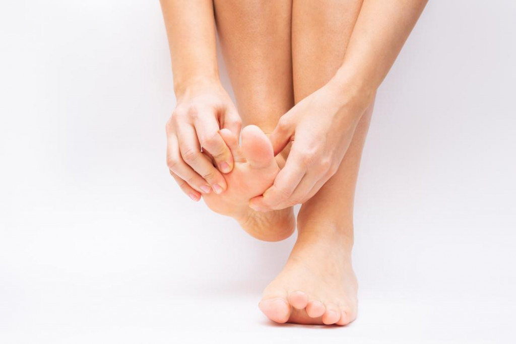 Pain in the foot and toes from uncomfortable shoes. A woman holds a sore foot with her hands on a white background. Pathology of bone structures, flat feet. Cramp, convulsion, spasm. Orthodontics