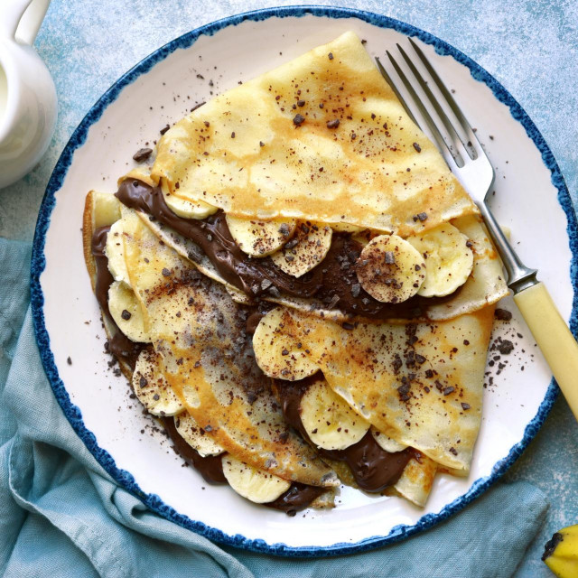 Thick pancakes with chocolate hazelnut spread and banana slices on a plate over light blue slate, stone or concrete background.Top view with copy space.
