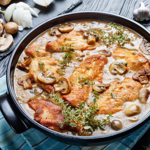 Chicken Marsala, Italian-American dish of golden pan-fried chicken cutlets and mushrooms in a rich Marsala wine sauce in a black ceramic dish on a wooden table, view from above, close-up