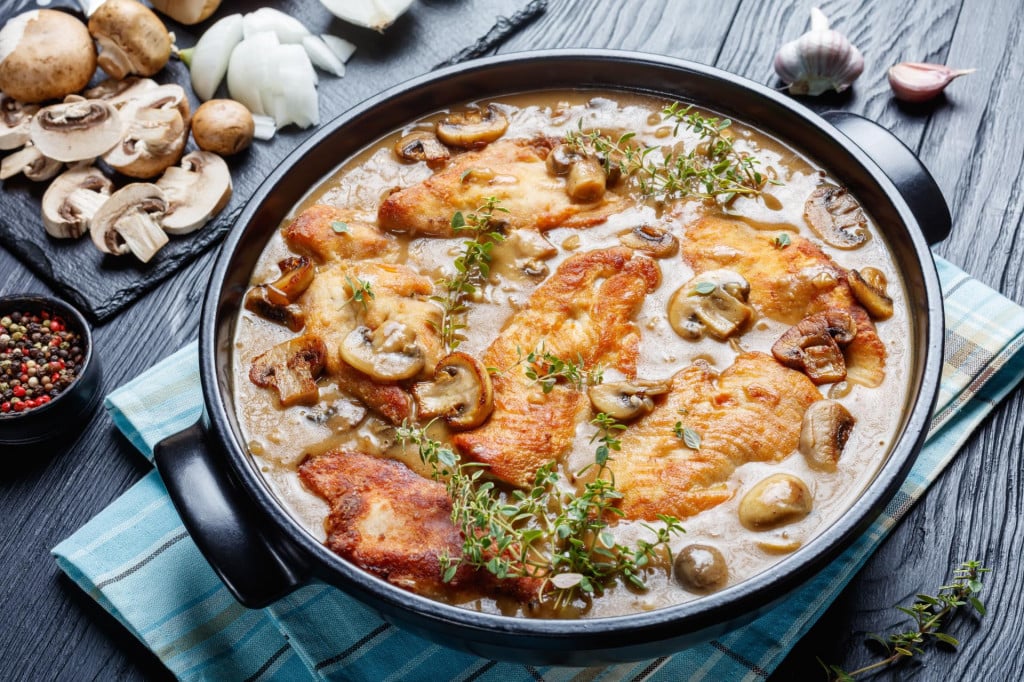 Chicken Marsala, Italian-American dish of golden pan-fried chicken cutlets and mushrooms in a rich Marsala wine sauce in a black ceramic dish on a wooden table, view from above, close-up