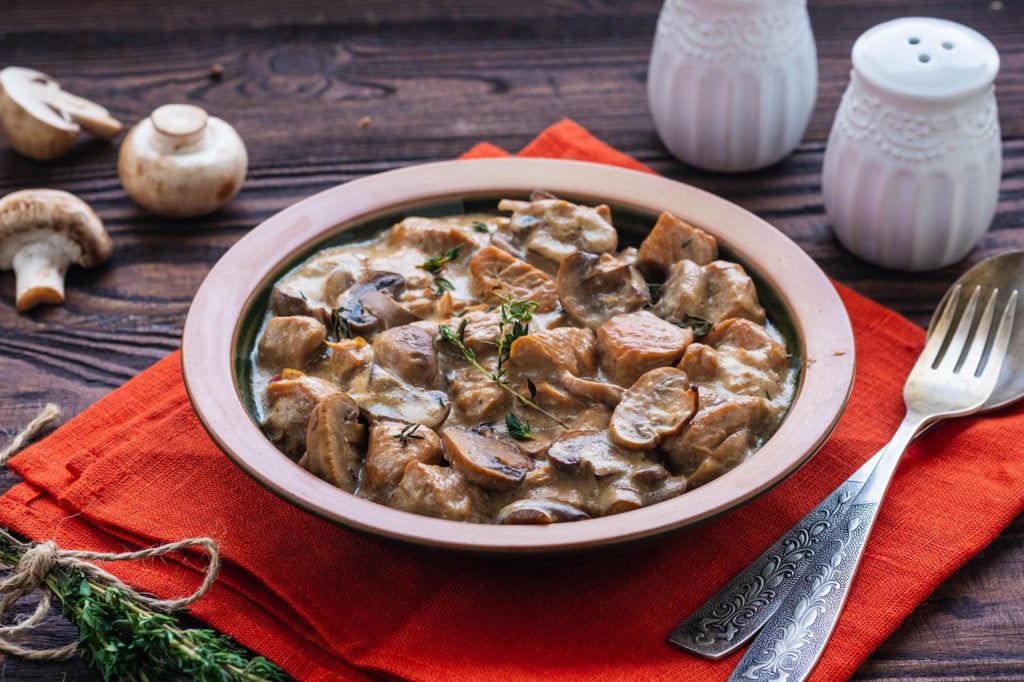 Stewed diced turkey with mushrooms in a creamy sauce in a clay plate on a brown wooden background. Turkey recipes. Second hot dishes