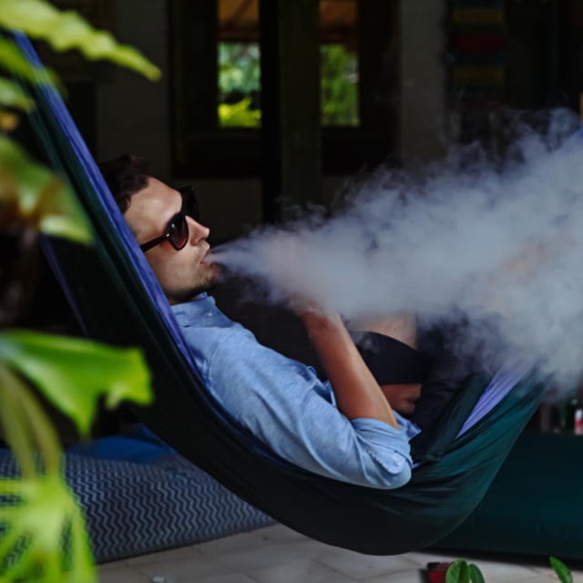 <p>Man resting in a hammock and vaping and letting off steam from an electronic cigarette</p>
