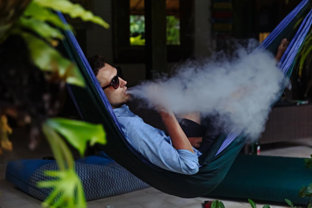 <p>Man resting in a hammock and vaping and letting off steam from an electronic cigarette</p>