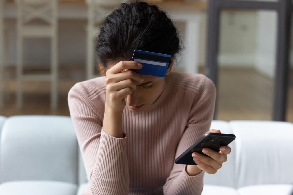 <p>Upset latina female internet shopper sit on couch hold phone with opened web shop ebank page suffer of overspending money from card account. Frustrated young lady lost savings as scam operation result</p>